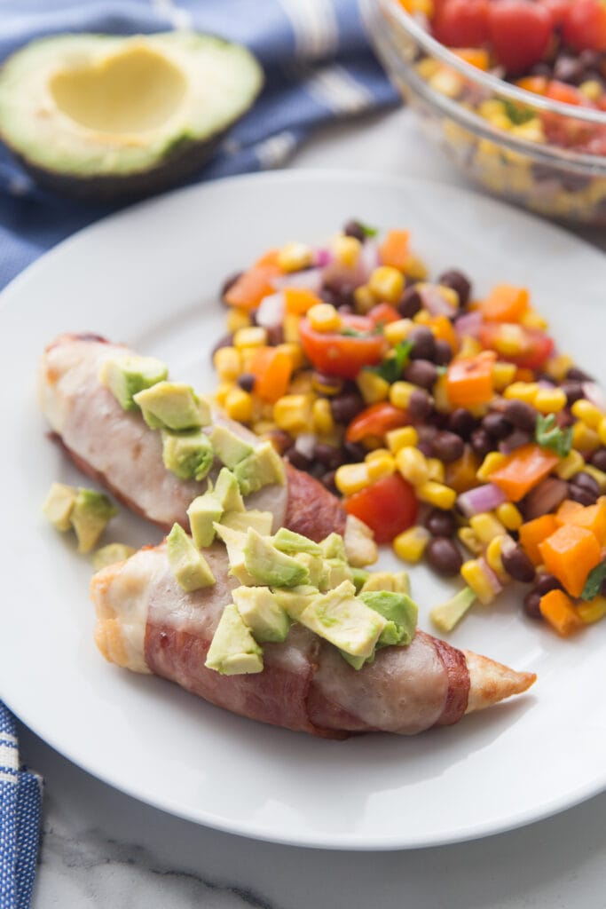A plate of bacon wrapped chicken sitting next to a corn and black bean salad
