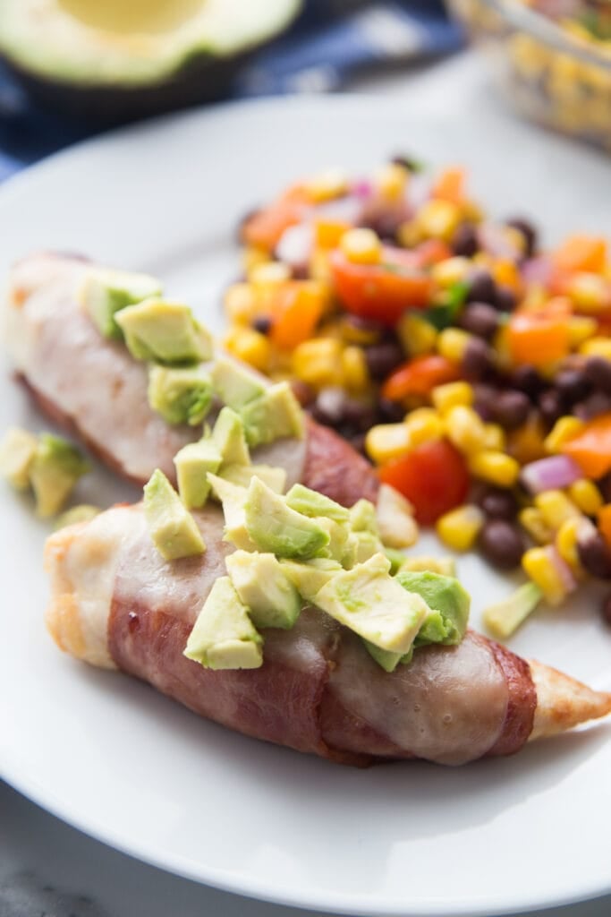 A plate with bacon wrapped chicken tenders topped with cheese and avocado sitting next to a black bean and corn salad 