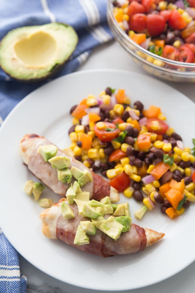 A overhead shot of air fryer bacon wrapped chicken tenders topped with cheese and avocado. A bowl of corn and black bean salad sits nearby and there is a half avocado sitting next to the plate on a kitchen towel. 