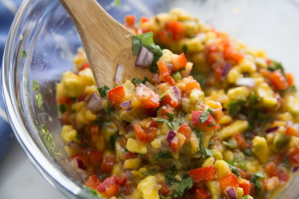 A bowl of avocado peach salsa being scooped up with a wooden spoon