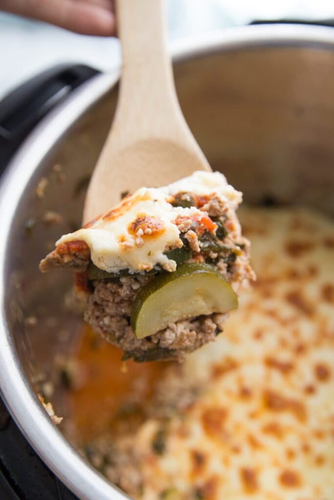 In the foreground- a wooden spoon with a bite of Instant Pot Zucchini Lasagna. In the background- (unfocused) an Instant Pot full of lasagna.