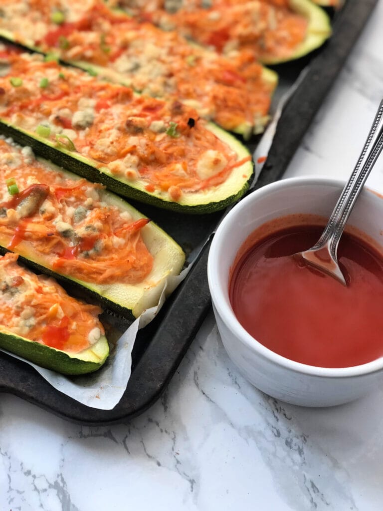 A tray of Buffalo Chicken Zucchini Boats next to a cup of hot sauce 