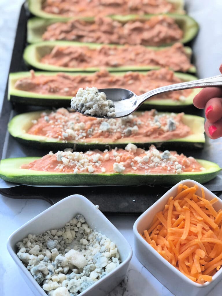 A tray of buffalo chicken stuffed zucchini being topped with blue cheese before baking
