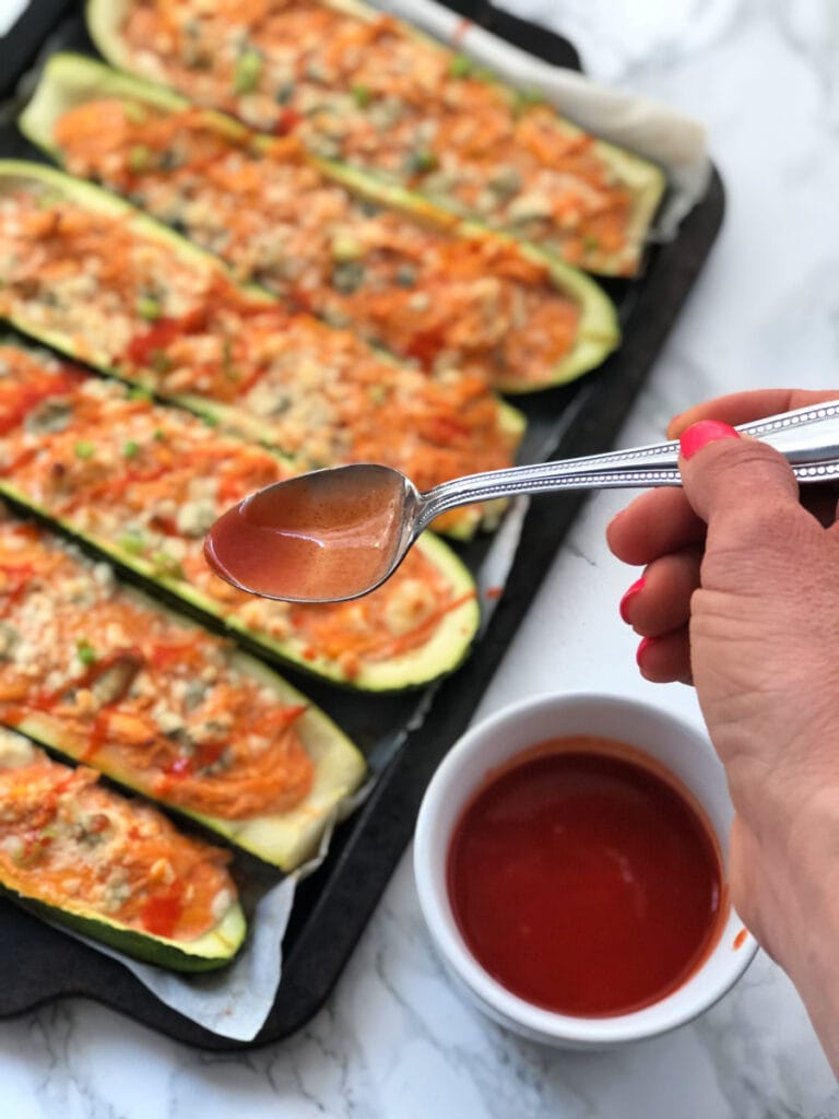 A spoon of hot sauce being drizzled over a pan of Buffalo Stuffed Zucchini Boats