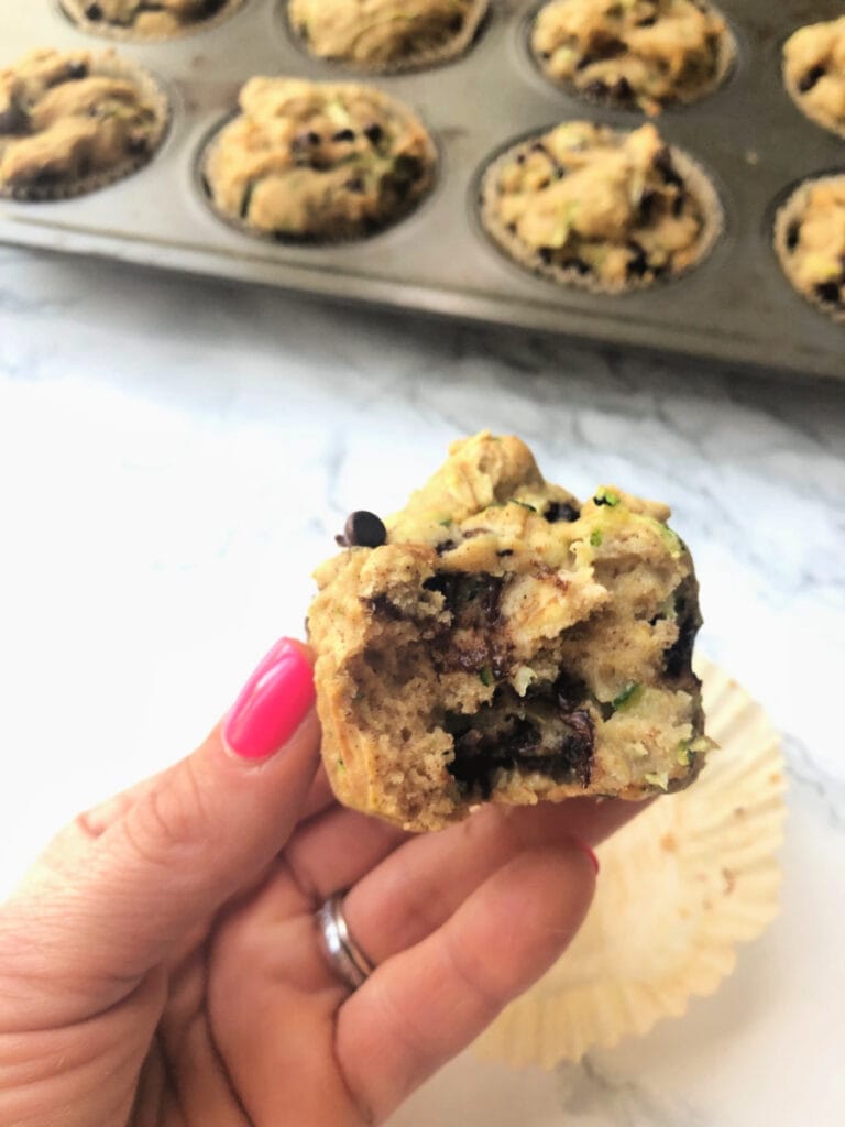 High angle of a woman's hand holding a chocolate chip zucchini muffin. The wrapper is behind the hand, on a white marble surface. In the background is a muffin tin full of more muffins.