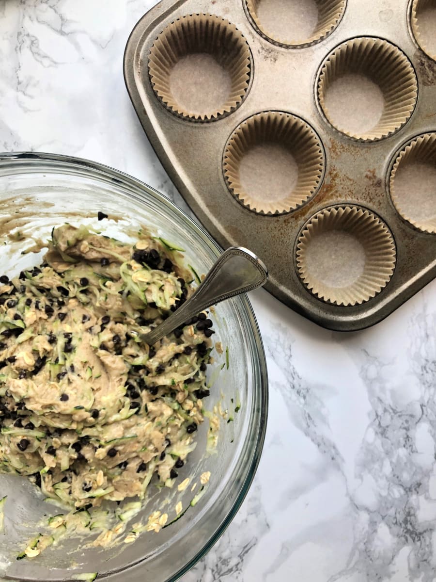 Overhead photo of Gluten free chocolate chip zucchini muffin batter in a glass bowl next to a muffin tin lined with parchment muffin liners.