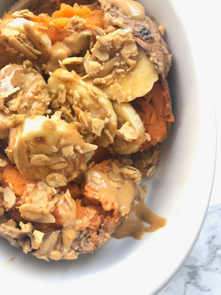 A close up shot of a sweet potato stuffed with banana, peanut butter and granola in a white bowl on a marble countertop