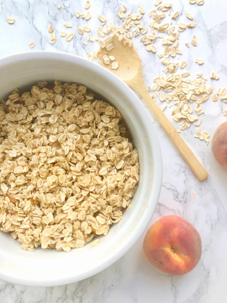 A casserole bowl of Instant Pot Peach Crisp sitting on a marble counter with a small wooden spoon and some peaches sitting nearby