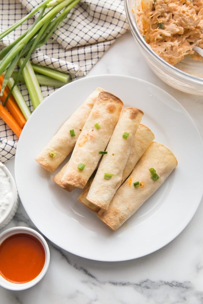 Gluten Free Buffalo Chicken Taquitos are stacked on a white dinner plate sprinkled with green onion. In the background is a glass bowl of extra buffalo chicken filling, ramekins of buffalo sauce and blue cheese dipping sauce, and celery and carrot sticks garnished with green onions..