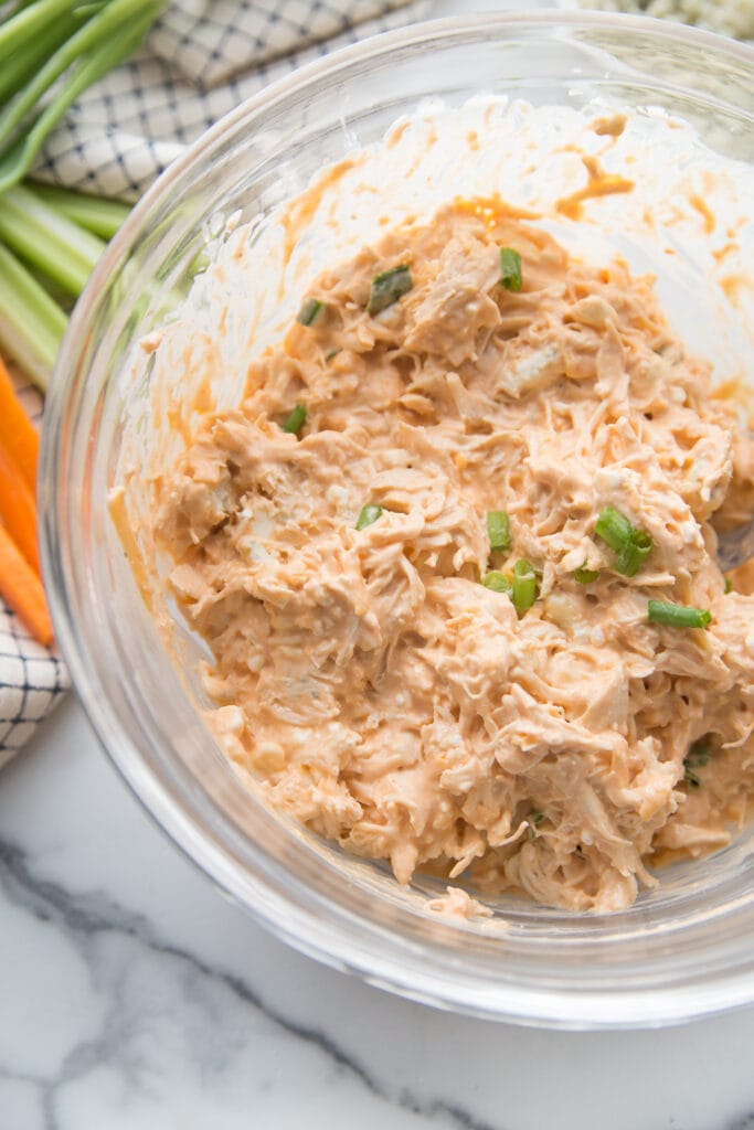A glass bowl of buffalo chicken taquito filling topped with sliced green onions and carrots and celery sticks in the background.