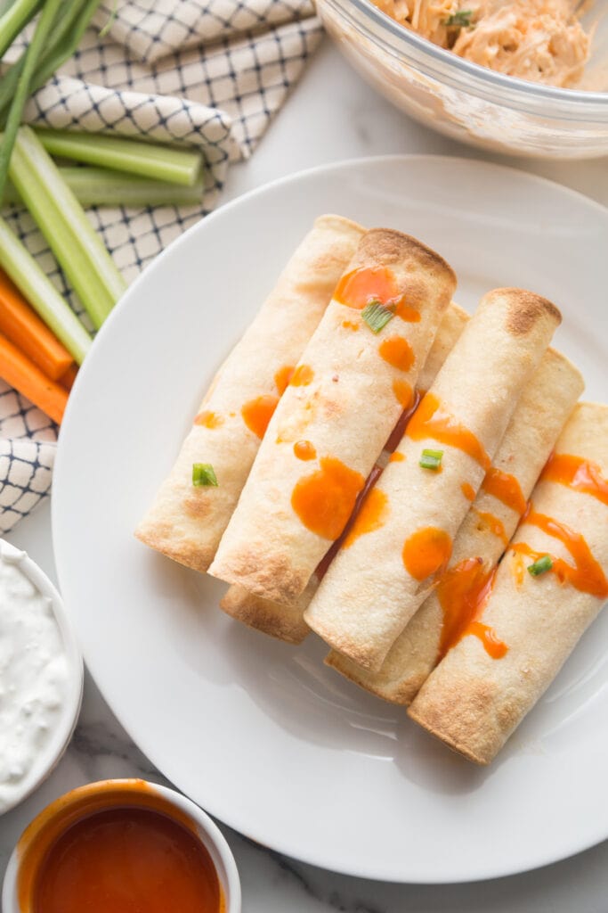 Gluten Free Buffalo Chicken Taquitos are stacked on a white dinner plate with buffalo sauce drizzled over top. In the background is a glass bowl of extra buffalo chicken filling, ramekins of buffalo sauce and blue cheese dipping sauce, and celery and carrot sticks.