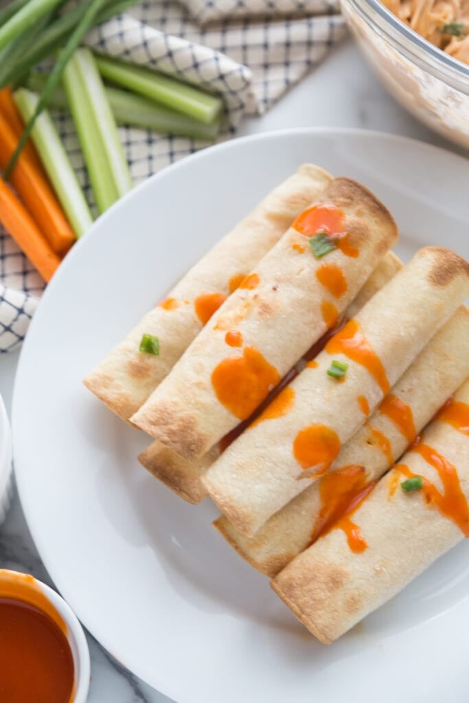 Gluten Free Buffalo Chicken Taquitos are stacked on a white dinner plate with buffalo sauce drizzled over top. In the background is a glass bowl of extra buffalo chicken filling, a ramekin of buffalo sauce, and celery and carrot sticks.
