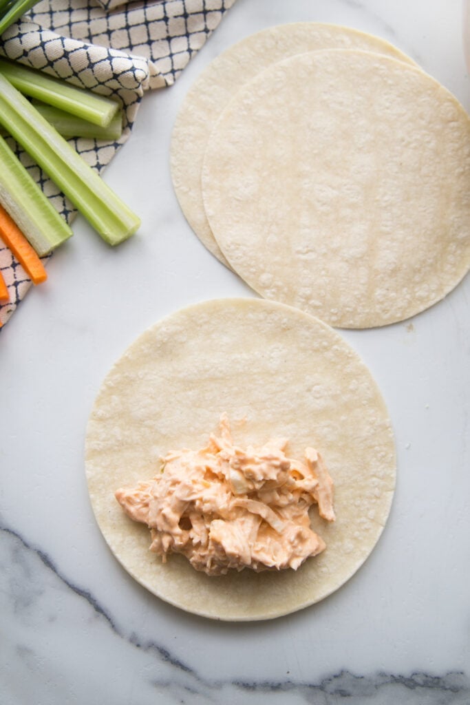 Shredded buffalo chicken is on a corn tortilla and ready to be rolled into a taquito. In the background are two more corn tortillas, carrot sticks and celery sticks on a checkered kitchen towel. 