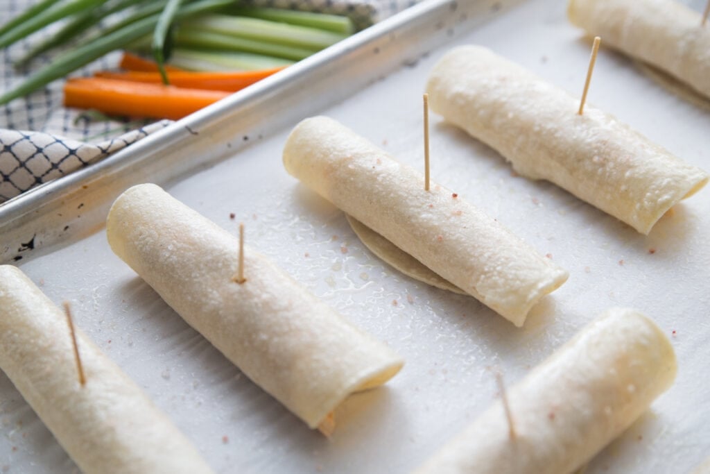 Buffalo chicken taquitos line a sheet pan ready to be put in the airfryer or oven. Each of the 6 taquitos have a toothpick securing them. In the background are celery sticks, carrot sticks, and green onions on a white and black plaid kitchen towel.