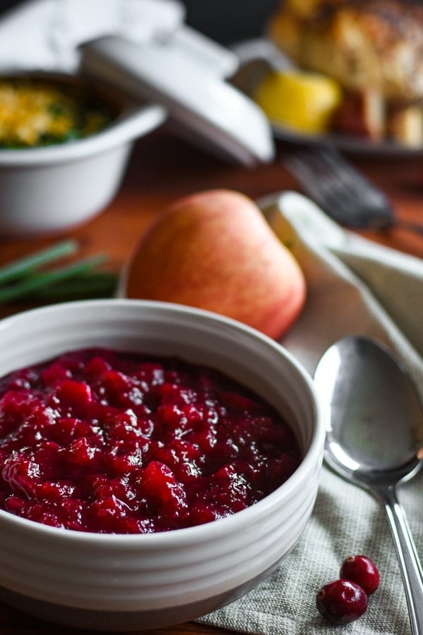 Close up photo of cranberry sauce in a small white bowl. In the background, out of focus, is a an apple and silverware, and a Thanksgiving table with a turkey and side dish. 