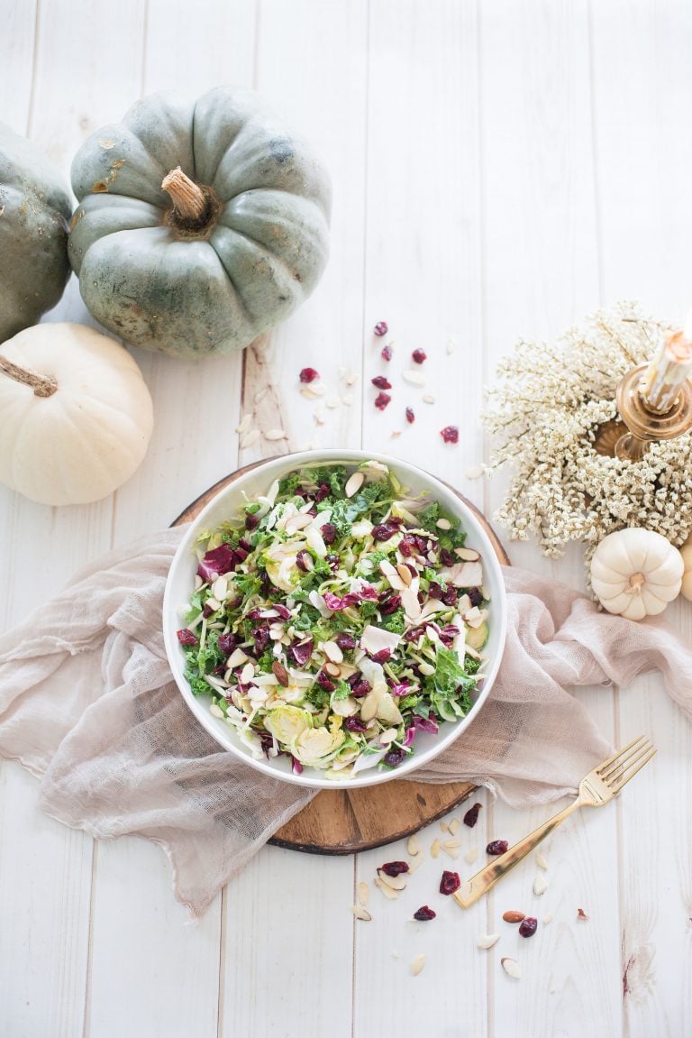 An overhead photo of Maple Cider Holiday Salad. The salad is in a white bowl and contains cabbage and greens, dried cranberries, and sliced almonds. The bowl is on a dark wooden circle with beige fabric underneath. There are white and blue/gray pumpkins, cranberries, almonds, and a fork decorating the white wooden surface.