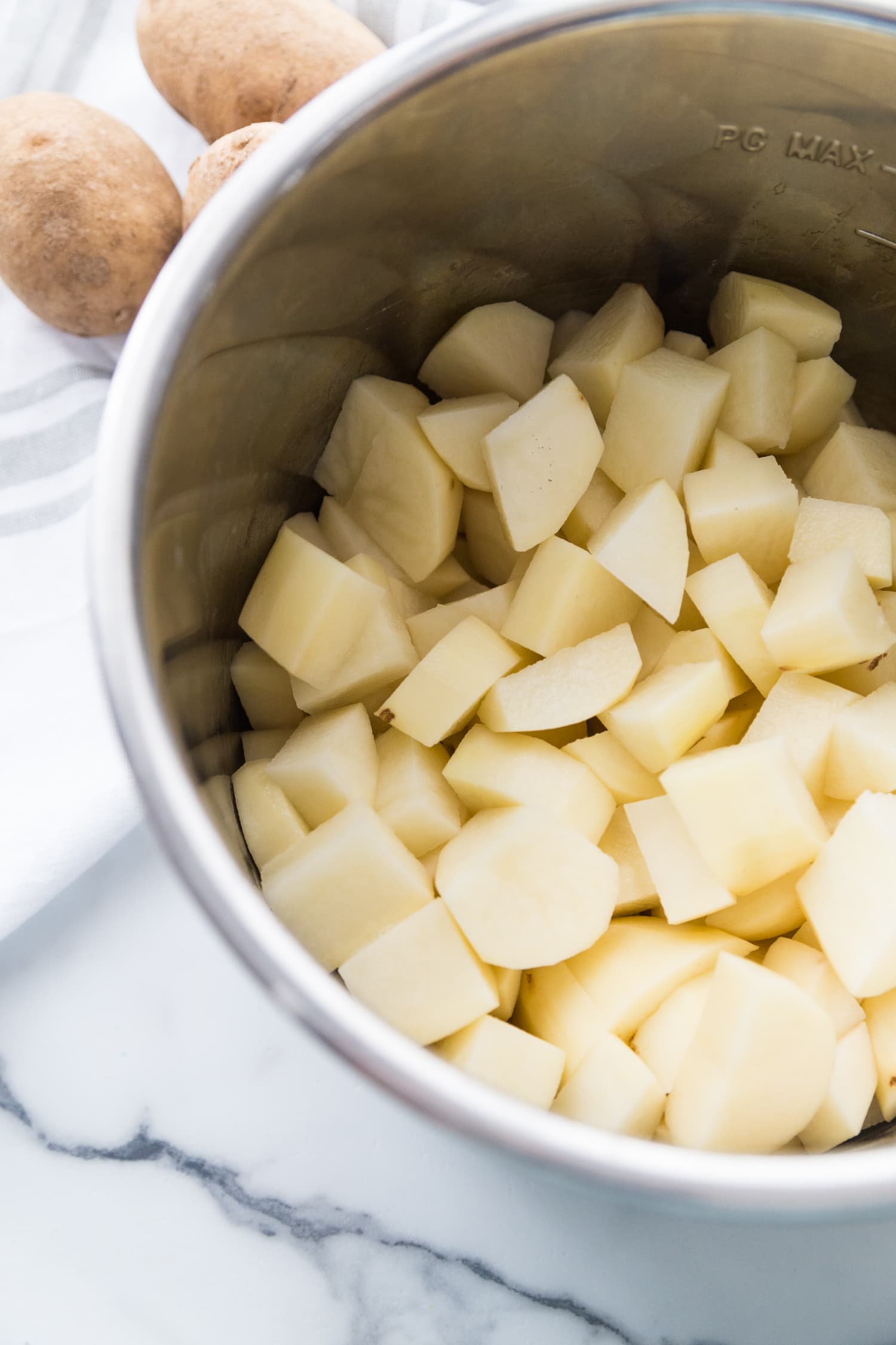 Instant pot full of peeled and diced potatoes on a white marble background with whole potatoes on the side.
