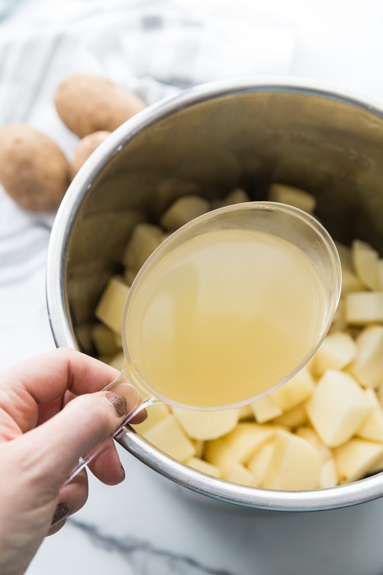 Overhead photo of peeled and diced potatoes in an Instant pot liner. Above the pot is a woman's hand holding a clear measuring cup full of chicken broth. 