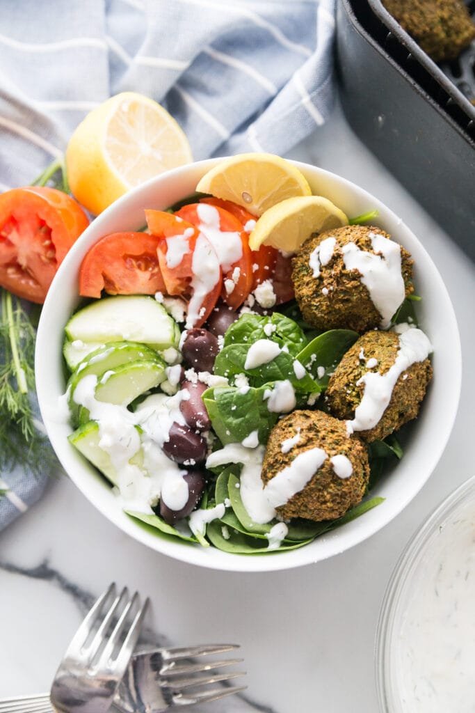 Overhead photo of large white bowl filled with Easy Falafel Bites and veggies including spinach, cucumbers, tomatoes, and olives, topped with tzatziki dressing with lemon wedges on the side. Off to the side are forks, an air fryer basket, a bowl of tzatziki dressing, veggies, and a blue kitchen towel.