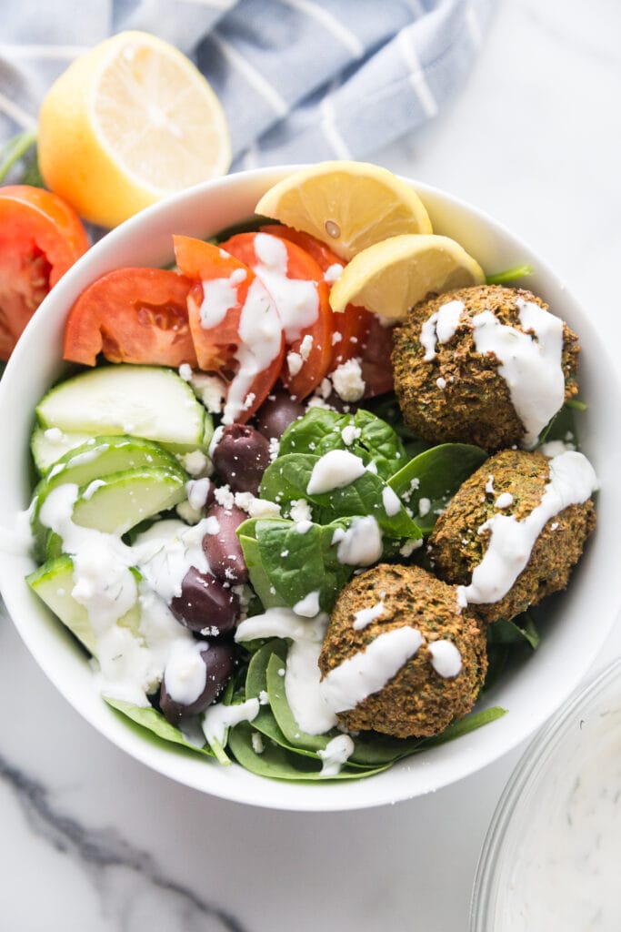 Overhead photo of large white bowl filled with Easy Falafel Bites and veggies including spinach, cucumbers, tomatoes, and olives, topped with tzatziki dressing with lemon wedges on the side.