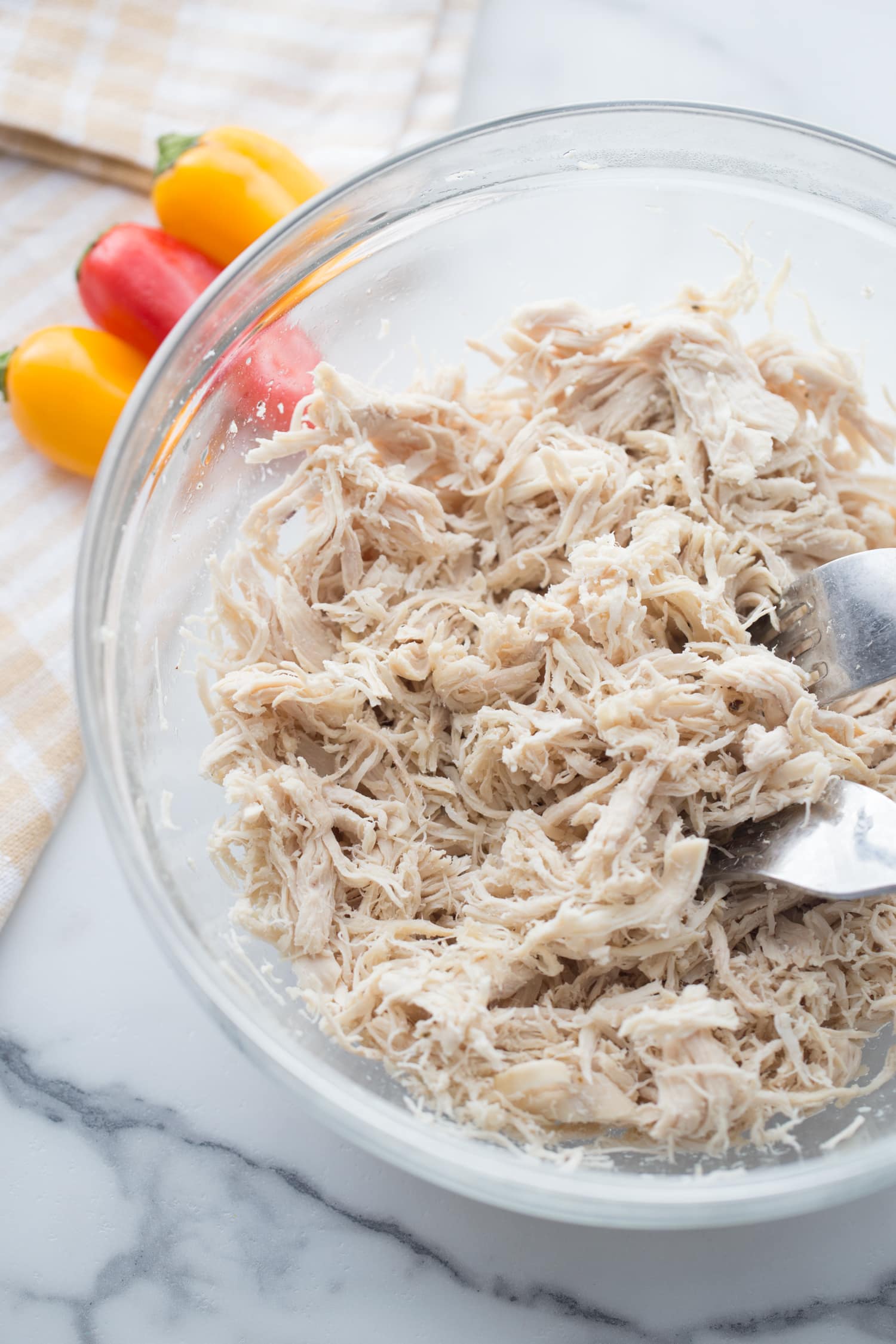 Shredded chicken in a glass bowl with two forks, mini bell peppers are in the background