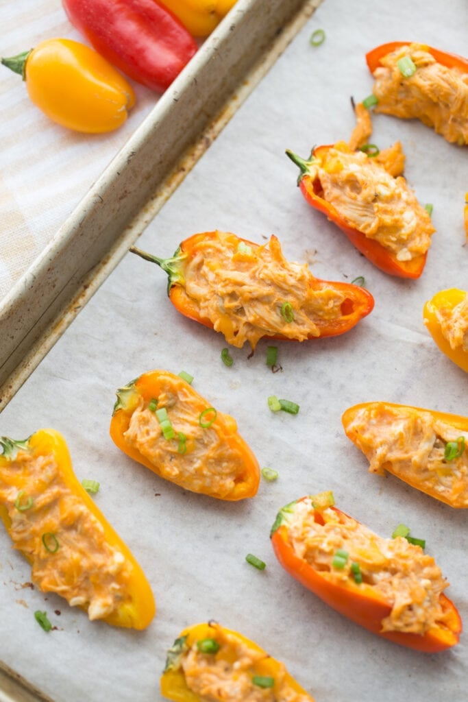 Close up of a sheet pan, turned diagonally, lined with parchment paper with rows of orange and yellow mini Healthy Buffalo Chicken Dip Stuffed Peppers