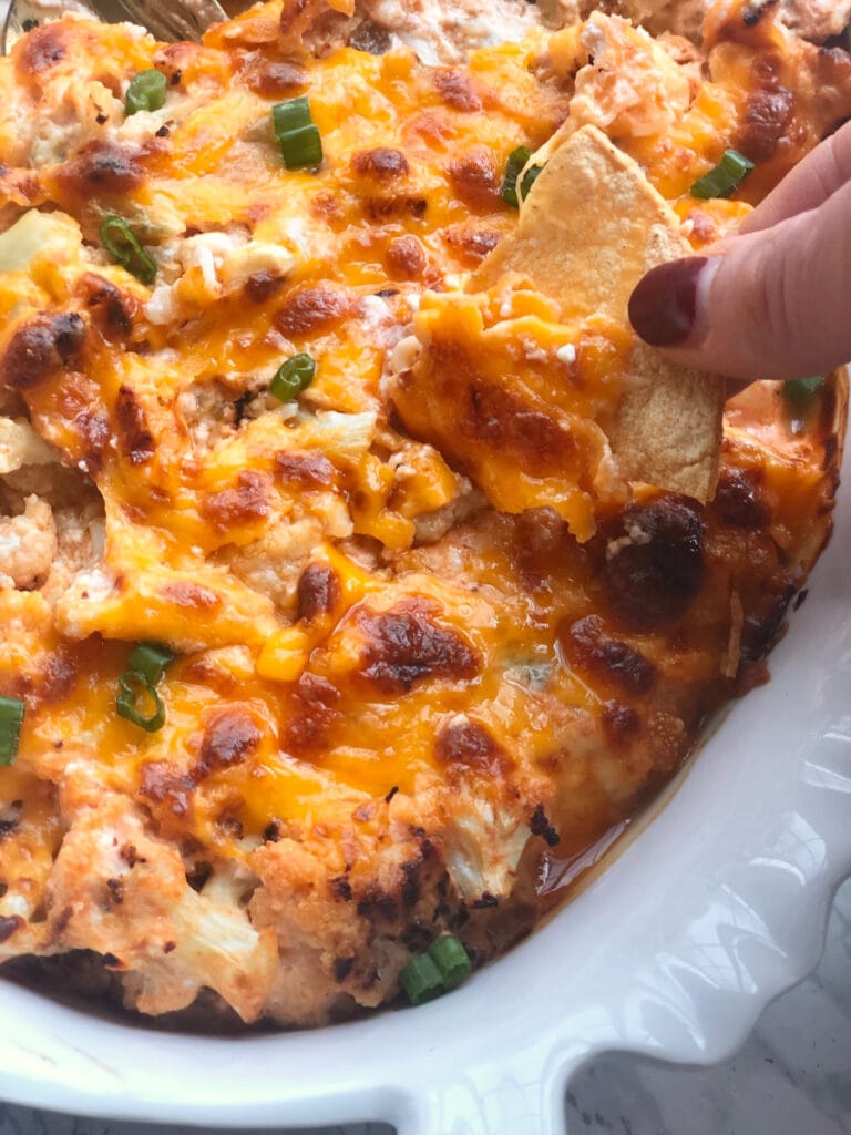 A woman's hand with painted fingernails is dipping a homemade tortilla chip into buffalo cauliflower dip. The dip is in a white oval serving dish with crispy, bubbly cheese on top and garnished with green onions- a great savory snack option.
