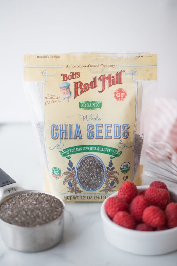A bag of Bob's Red Mill Chia Seeds in the center with a shallow white ramekin full of whole raspberries and a measuring cup full of chia seeds in front of the bag.