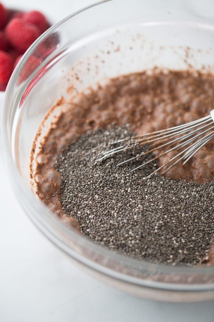 Chocolate pudding in a large glass bowl with raw chia seeds on top. A whisk is in the pudding, ready to mix in the chia seeds.