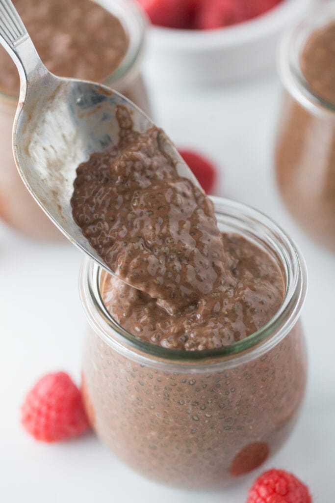 Silver spoon with a bite of chocolate chia pudding rests over the small individual glass container of pudding. Raspberries are on the side.