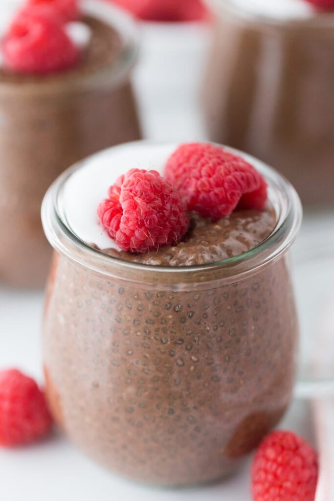 Close up photot of Chocolate chia pudding in a glass cup topped with whole raspberries and coconut cream. There are more raspberries and glass containers full of chocolate chia pudding in the background, out of focus.