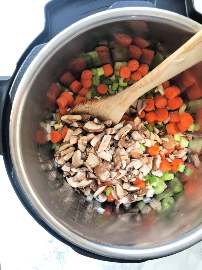 Overhead view of an instant pot full of chopped veggies- carrots, celery, onion, mushrooms- with a wooden spoon, ready to stir.