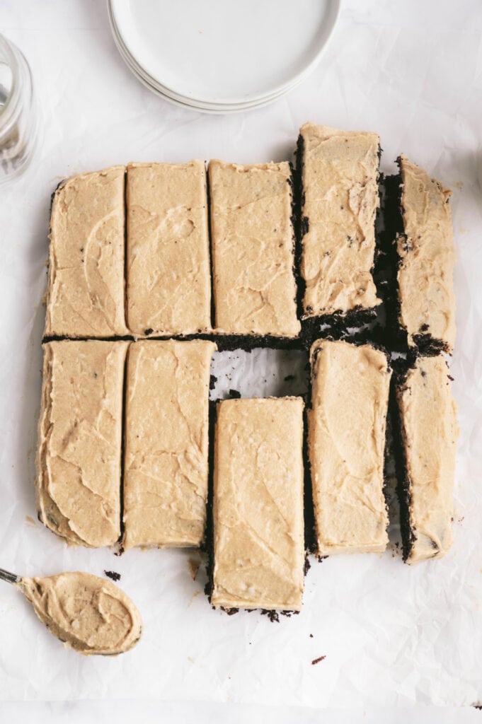 Overhead photo of Chocolate cake with peanut butter frosting cut into 10 rectangular pieces. There is a spoonful of frosting off to the side as well as round white plates.