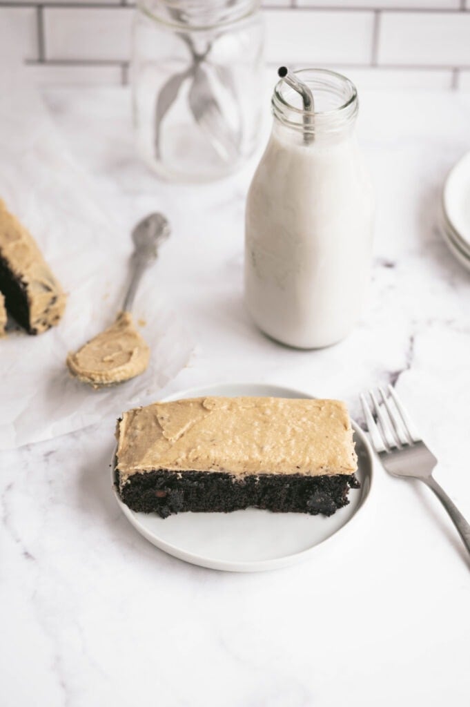 Slightly angled overhead photo of a piece of chocolate cake on a white plate. It has a thick layer of peanut butter frosting on top. There is a glass of milk and a fork on the side, ready to dig in.