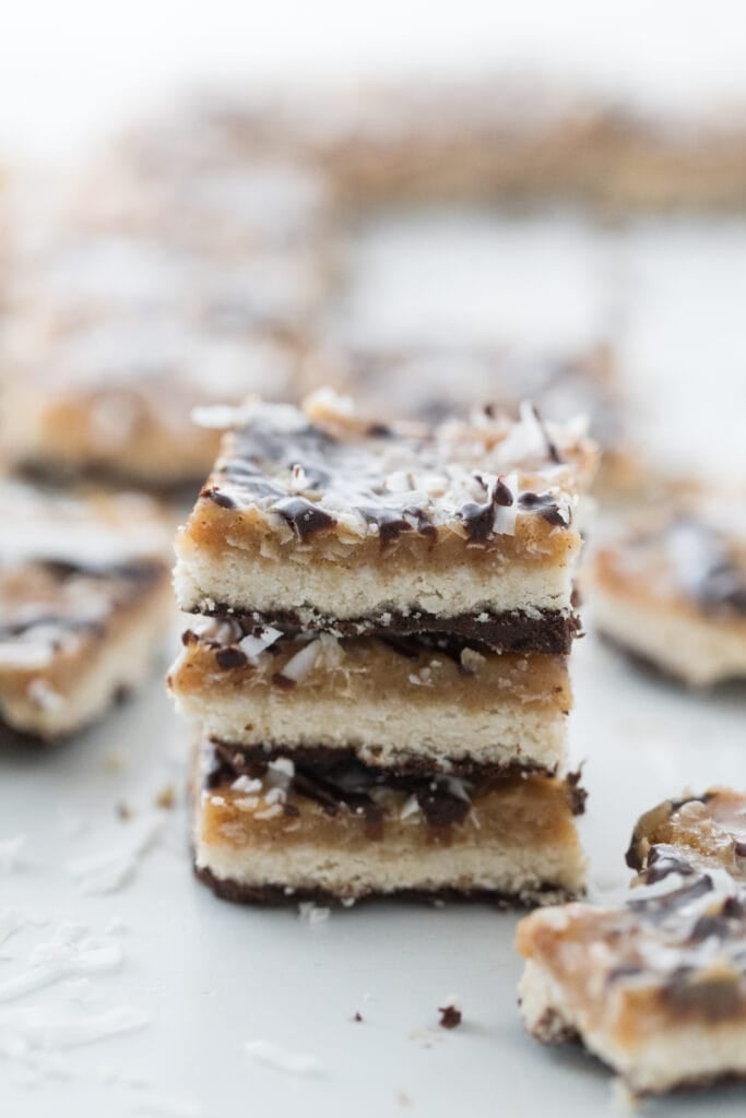 Healthy Samoas Cookie Bars stacked on top of one another showing the layers of coconut, homemade caramel, and chocolate; more cookie bars are scattered in the background, out of focus