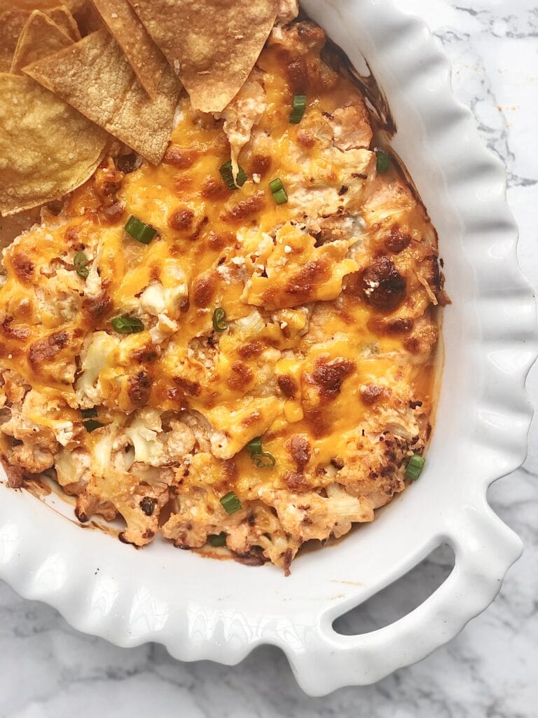 Overhead photo of Buffalo Cauliflower Dip topped with browned, crispy cheese in a white oval casserole dish with homemade tortillas on the side.