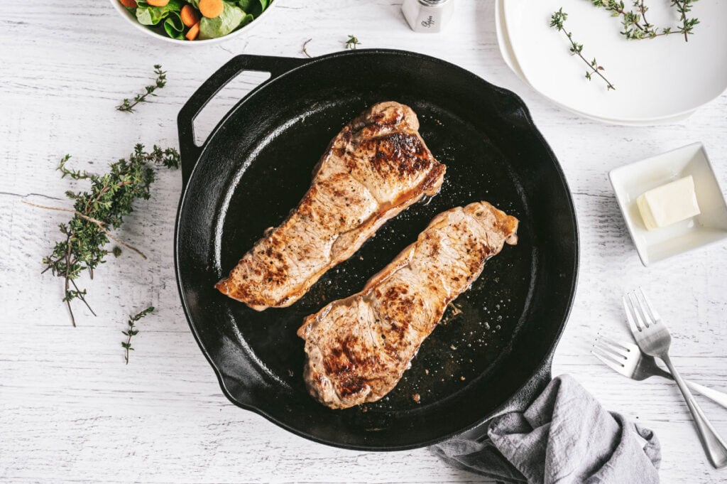 Overhead photo of New York Strip Steaks grilled in a cast iron skillet with thyme, butter, forks, and salad on the side.