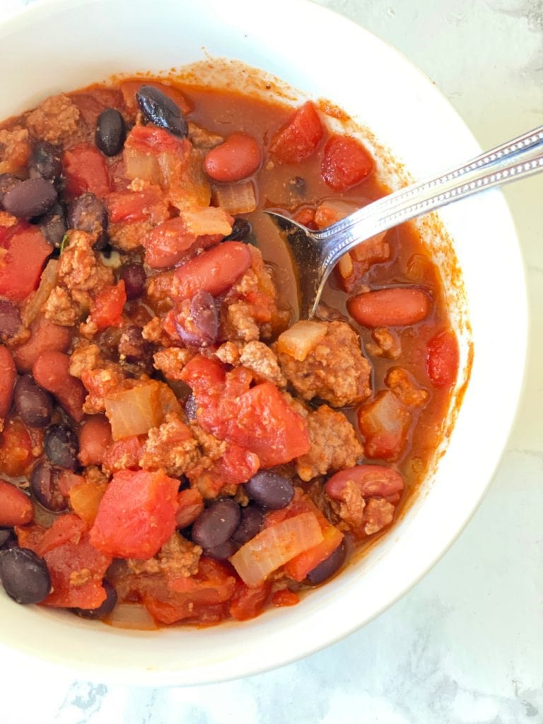 Overhead close up photo of 5 ingredient chili, made with pantry ingredients like beans, diced tomatoes, and onions in a white bowl with a spoon on the side.