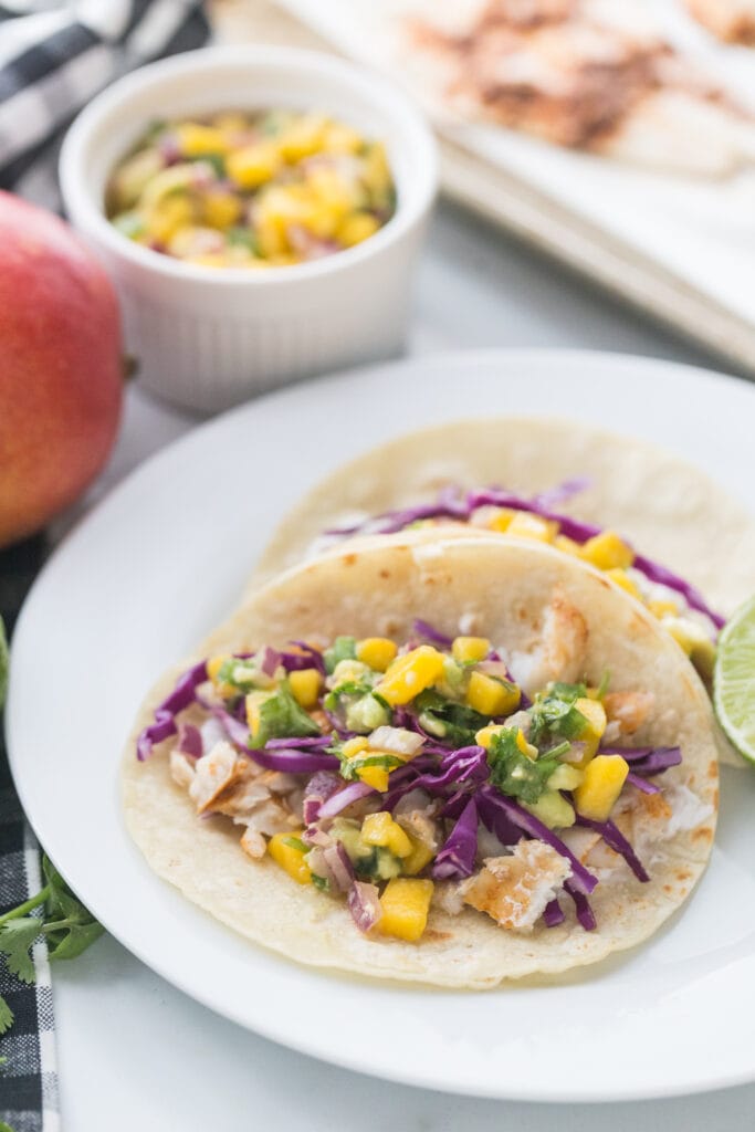 Side view of Two Fish Tacos with Mango Salsa and red cabbage on corn tortillas on a white plate. In the background, out of focus is a whole mango, extra salsa in a ramekin, and a pan of fish.