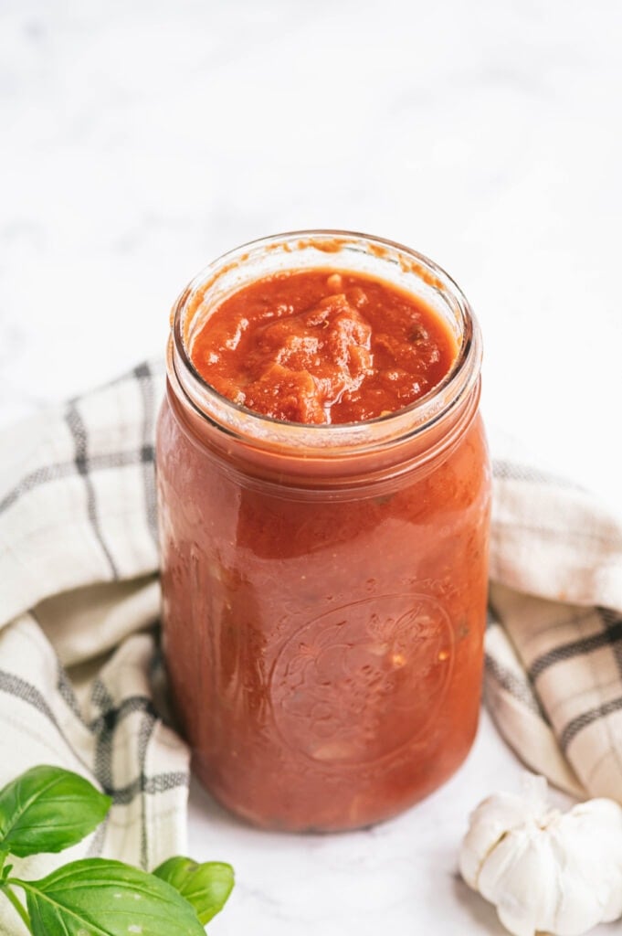 High angle of a large mason jar full to the brim of homemade tomato sauce. Near the jar is a white and blue plaid towel, some fresh basil, and a bulb of garlic on a white surface.
