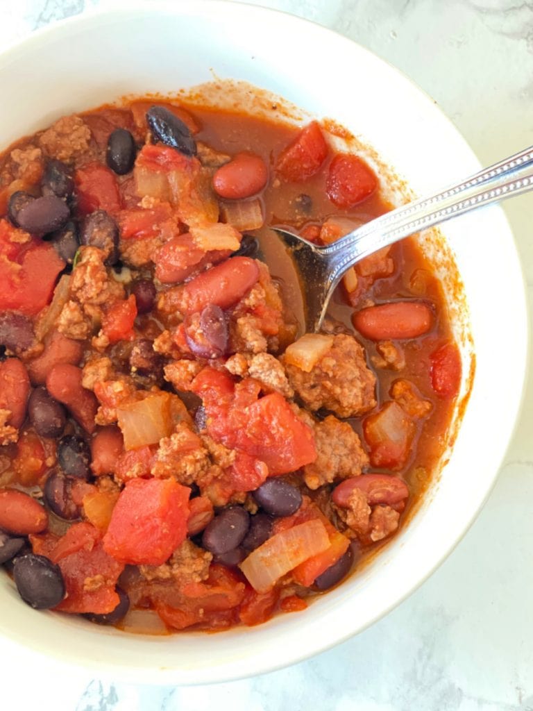 Close up photo of chili made mostly with pantry ingredients- canned tomatoes, black beans, kidney beans, ground meat, onions- in a white bowl with a spoon