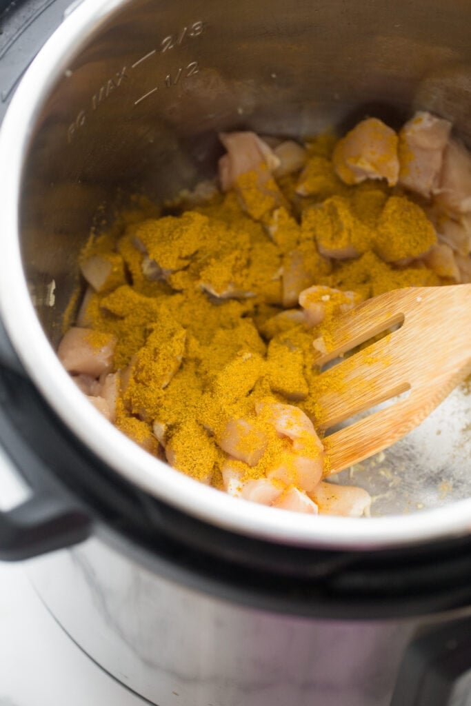Overhead view of diced raw chicken, covered in yellow curry seasoning, in an Instant Pot with a slotted wooden utensil. 