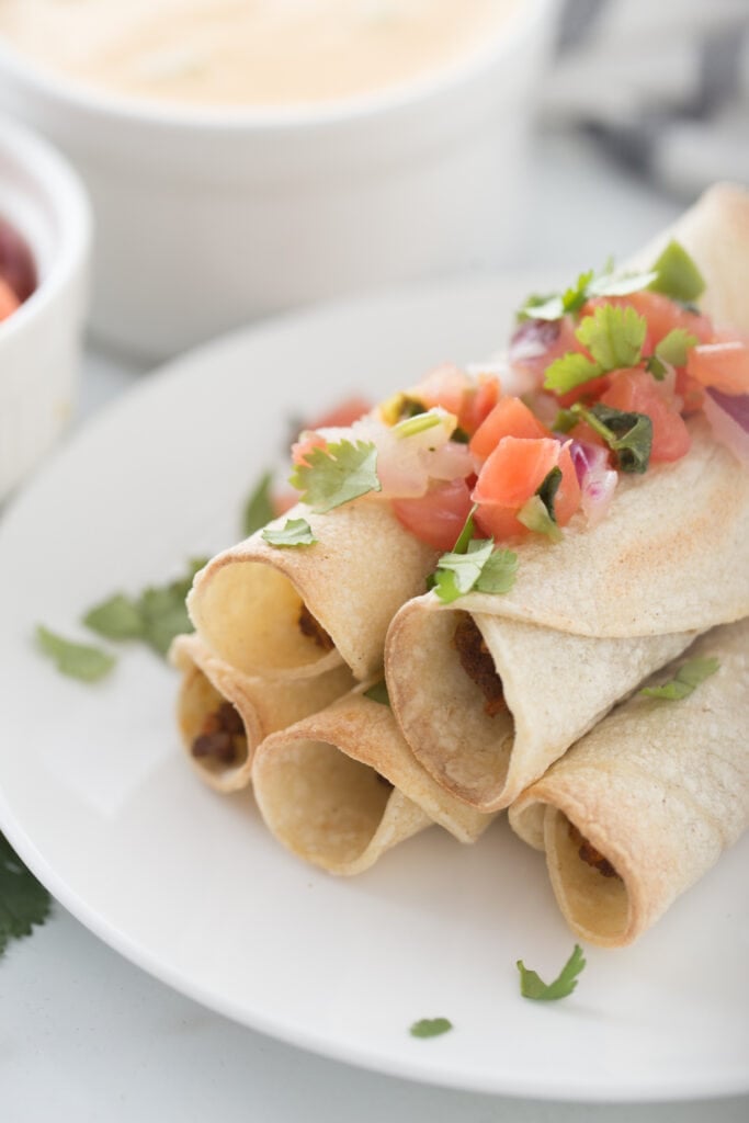 Close up photo of a plate stacked with beef taquitos.topped with homemade salsa. In the background, out of focus, are ramekins filled with salsa and cheese sauce.