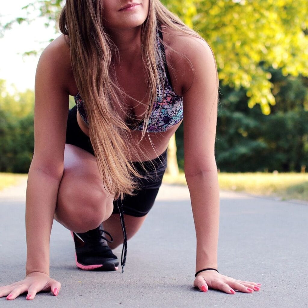 A medium-blonde, long haired woman wearing black shorts, multicolored sports bra, and black and pink sneakers is in a runner's lunge. She is outside on a path with green trees in the background. 