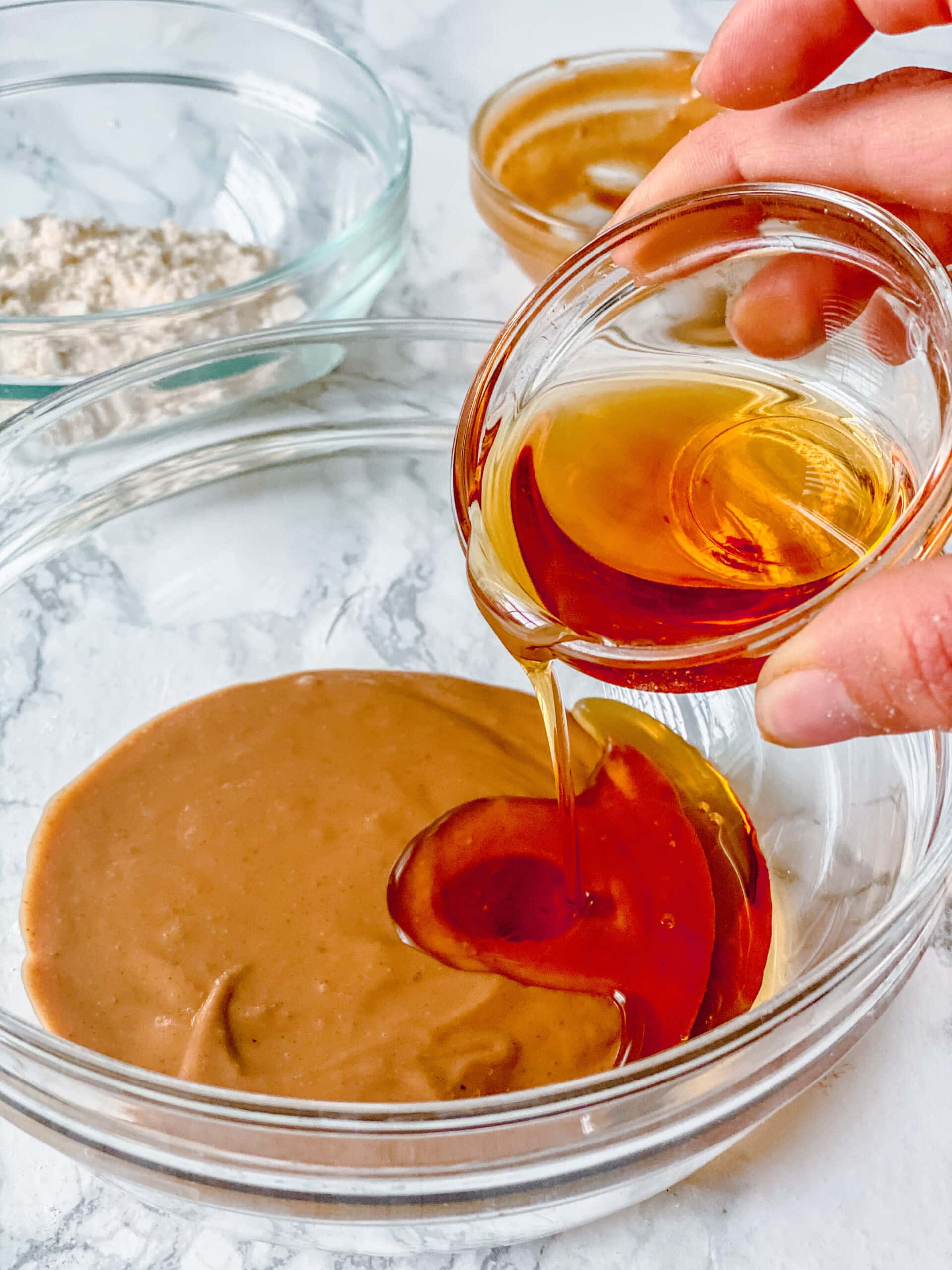 Assorted glass bowls filled with ingredients to make simple peanut butter cookies. A hand is holding the smallest bowl and pouring maple syrup into the larger bowl full of peanut butter.