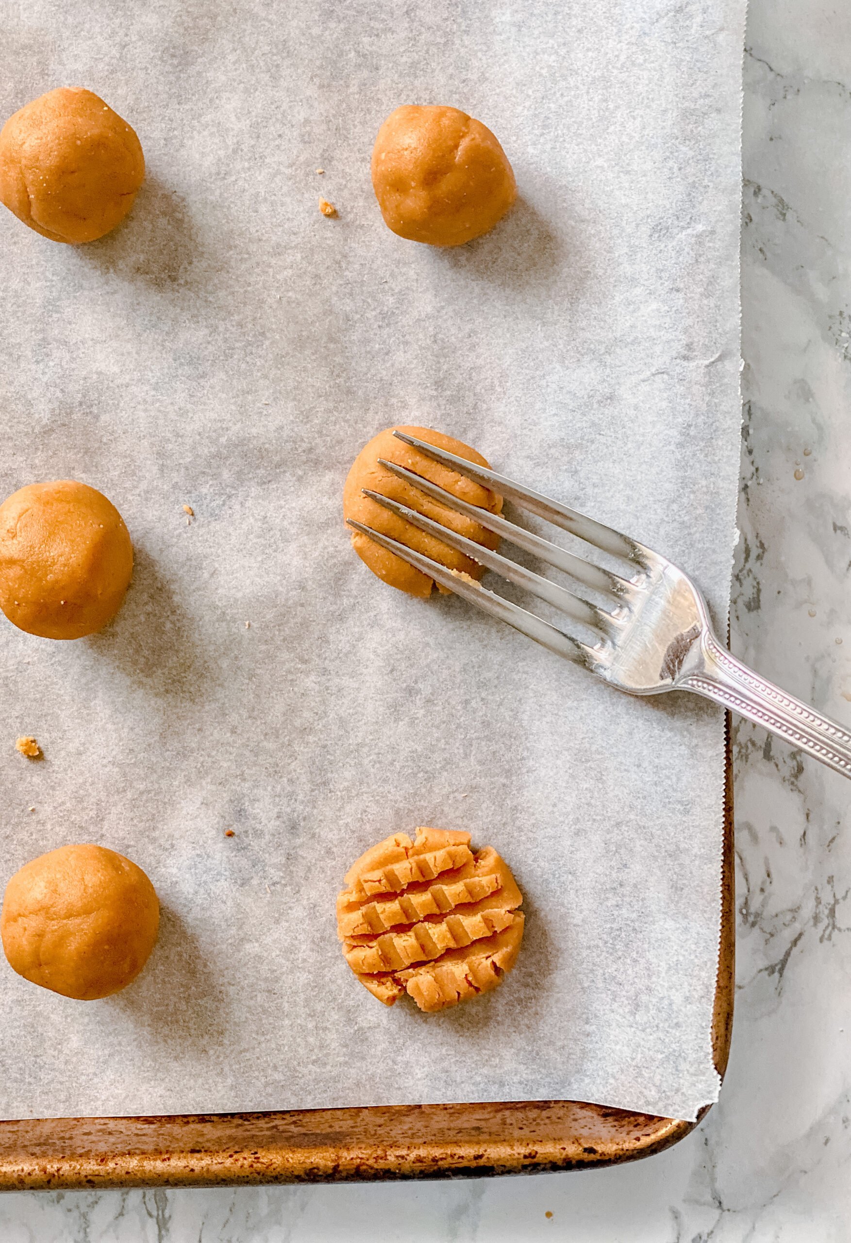 Parchment lined baking sheet with balls of no bake peanut butter cookie dough. One has been flattened with a fork to make a criss cross pattern, one has a fork in it, creating the texture.