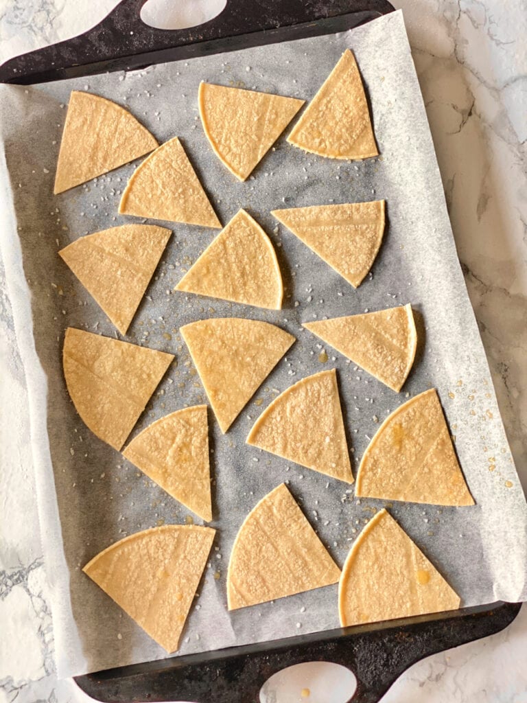 Overhead photo of homemade tortilla chips made out of corn tortillas cut into triangles. The chips are on a parchment lined sheet pan on a marble surface.