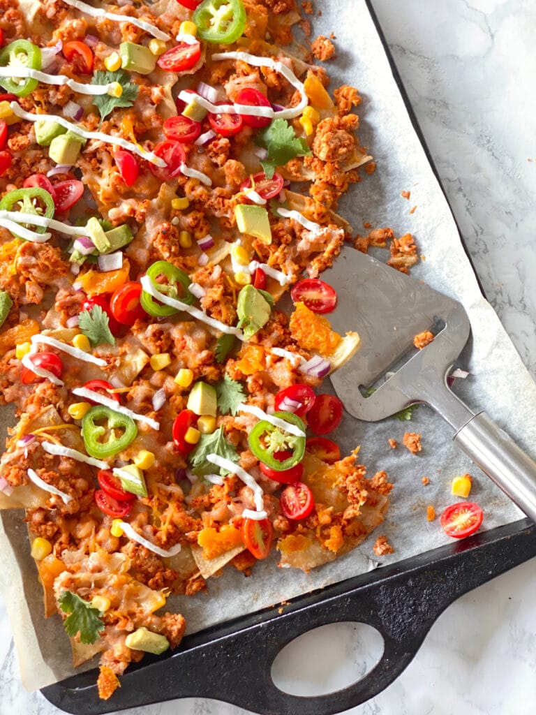 Homemade nachos topped with ground beef, cheese, jalapeno, tomatoes, corn, cilantro, avocado, and drizzled Greek yogurt on a parchment lined sheet pan. In the corner of the sheet pan, some of the nachos have been served and there is a silver utensil resting on the pan.