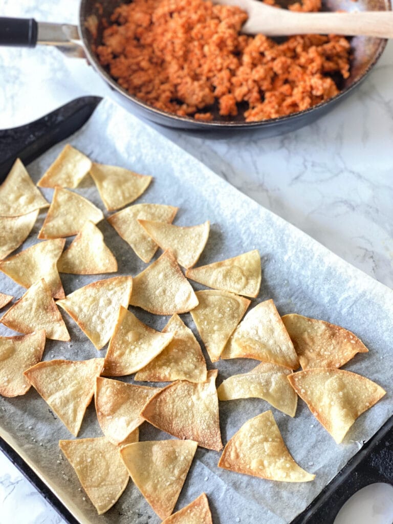 Cooked homemade tortilla chips with browned crispy edges on a parchment lined sheet pan. At the top of the photo is a skillet of cooked ground beef with a wooden spoon,