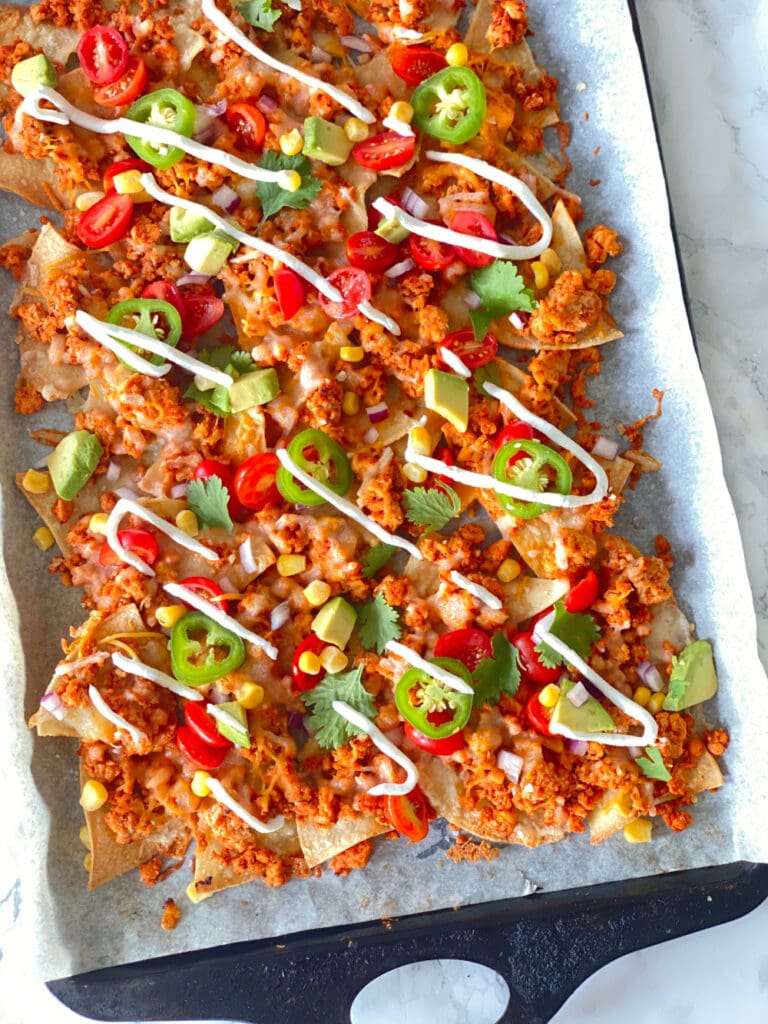 Homemade nachos topped with ground beef, cheese, jalapeno, tomatoes, corn, cilantro, avocado, and drizzled Greek yogurt on a parchment lined sheet pan.